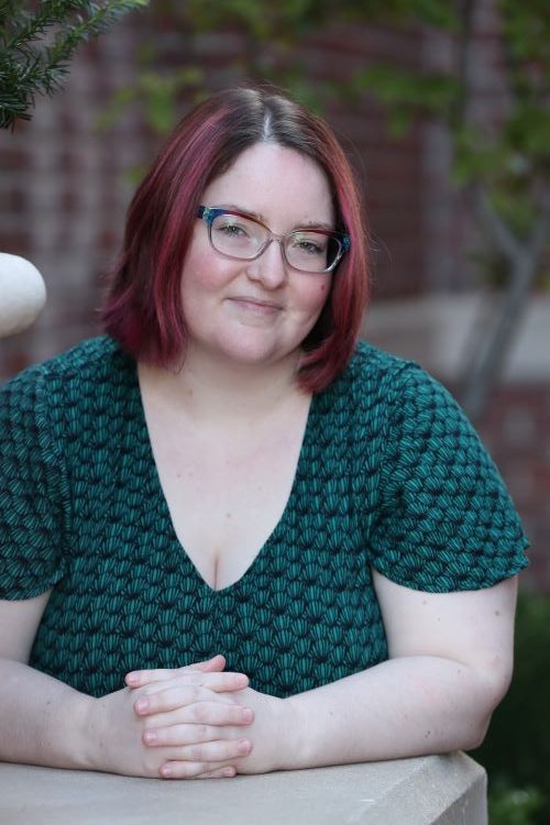 The author, Shimy Moesel, a white person with brown and pink hair and rainbow watercolor glasses, smiling at the viewer from another angle.
